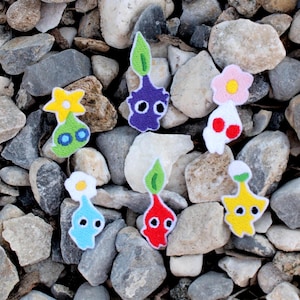 Puede incluir: Una colección de coloridos personajes Pikmin de fieltro, cada uno con diseños y características únicas, están dispuestos sobre un lecho de piedras grises y beige. Los Pikmin tienen varios colores, incluyendo rojo, azul, amarillo, morado y blanco, con copas en forma de flor u hoja.