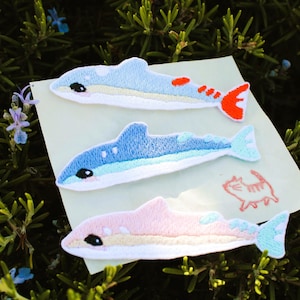 May include: Three embroidered iron-on patches shaped like dolphins, arranged vertically on a light-colored card. The top patch is light blue with orange accents, the middle is blue, and the bottom is pink. A small red line drawing of a cat is visible.