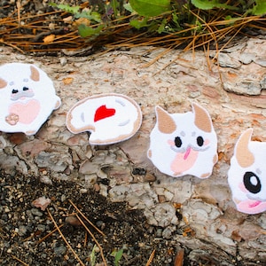 May include: Four embroidered patches featuring three white, horned creature designs with pink accents and large black eyes, alongside one white, oval-shaped patch with a red heart in the centre, all displayed on a textured, light brown wooden surface.