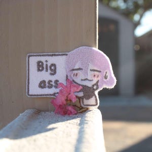 Puede incluir: Un parche de tela con un personaje de estilo anime con cabello lavanda y un atuendo negro. El parche incluye un letrero que dice "Big ass" en fuente blanca. Flores rosas se colocan cerca del personaje.