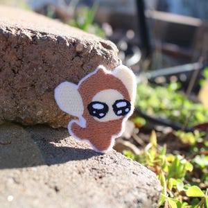 Puede incluir: Un parche bordado marrón y blanco que representa una criatura de dibujos animados con ojos grandes y expresivos. El parche está colocado sobre una superficie de piedra rugosa, con vegetación en el fondo. El diseño es caprichoso y adecuado para ropa o accesorios.