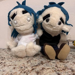 May include: Two plush dolls with cream-colored fur, blue hair, and thick black eyebrows. One doll wears a white t-shirt, while the other wears a black top and dark pants with brown feet. Both dolls have white circular eyes and sit side by side.