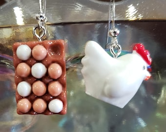 Pode incluir: Brincos com um design de caixa de ovos castanha com ovos brancos e castanhos, e uma galinha branca com crista e bico vermelhos. Os brincos penduram-se de ganchos prateados.