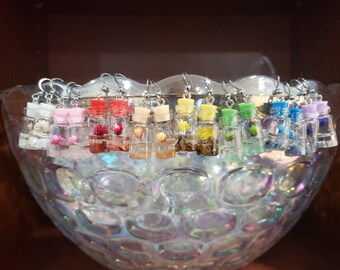 Könnte beinhalten: Eine Regenbogen-Auswahl an kleinen Glasflaschen, gefüllt mit getrockneten Blumen und in klarem Harz gegossen. Jede Flasche ist an einem silbernen Ohrringhaken befestigt. Die Ohrringe werden in einer durchsichtigen Glasschale präsentiert.