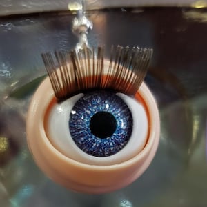 May include: A close-up of a doll eye with a blue iris and brown eyelashes. The eye is set in a light peach-colored socket. The eye is suspended by a silver wire.
