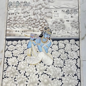 May include: A detailed painting depicts a blue-skinned figure playing a flute, seated amidst white lotus flowers. The background shows a pastoral scene with trees, cows, and a gray sky. The artwork is framed with decorative wooden stamps at the corners.