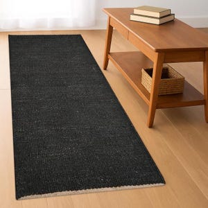 Puede incluir: Una alfombra negra rectangular y larga con una superficie texturizada y un borde color crema. La alfombra está colocada sobre un suelo de madera clara. Una mesa de centro de madera con libros y una cesta de mimbre está en el fondo.