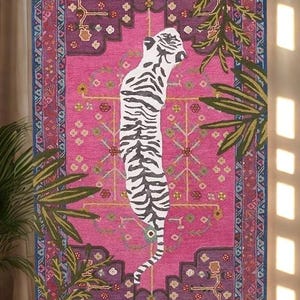 May include: A rectangular rug featuring a pink central field with a white and black zebra design. The rug is framed by a decorative border in purple and blue hues, with green palm leaves on the sides.