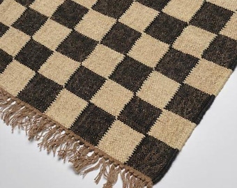May include: A woven rug featuring a black and beige chequerboard design. The rug has a fringed edge in a light brown hue. The rug is constructed from a natural fibre material.