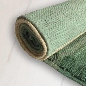 Puede incluir: Alfombra verde enrollada con un tejido texturizado y un borde beige. La alfombra tiene un detalle de flecos en el borde, y la paleta de colores incluye tonos de verde y beige. La alfombra probablemente está hecha de fibras naturales.
