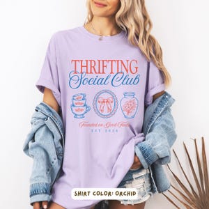 May include: Orchid-colored t-shirt with the text "THRIFTING Social Club" in red and blue. The design includes illustrations of teacups, a bow, and a vase with flowers. The text "Founded on Good Finds" and "EST 2026" are also present.