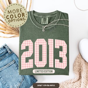 May include: Moss green t-shirt with the year "2013" in pink gingham print. The shirt has the words "Limited Edition" printed below the numbers. A silver necklace with a pendant is on the shirt. The words "More Color Options" are in a circle.