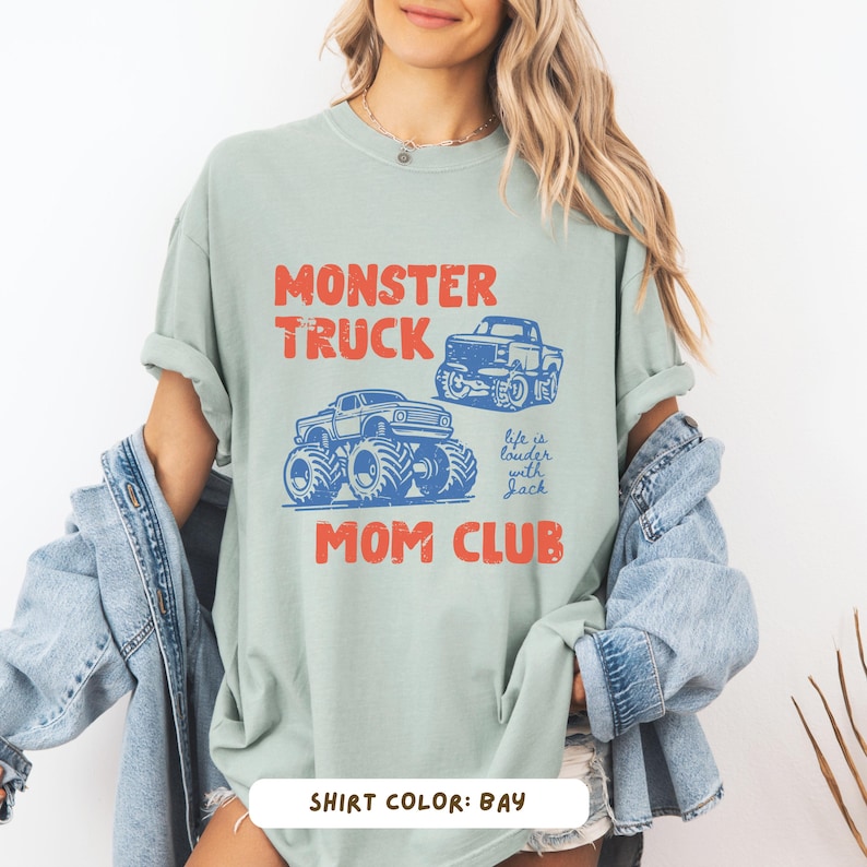 May include: A light green t-shirt with the words "MONSTER TRUCK MOM CLUB" in red and blue. The shirt features two monster truck illustrations and the text "life is louder with Jack". The shirt color is bay.