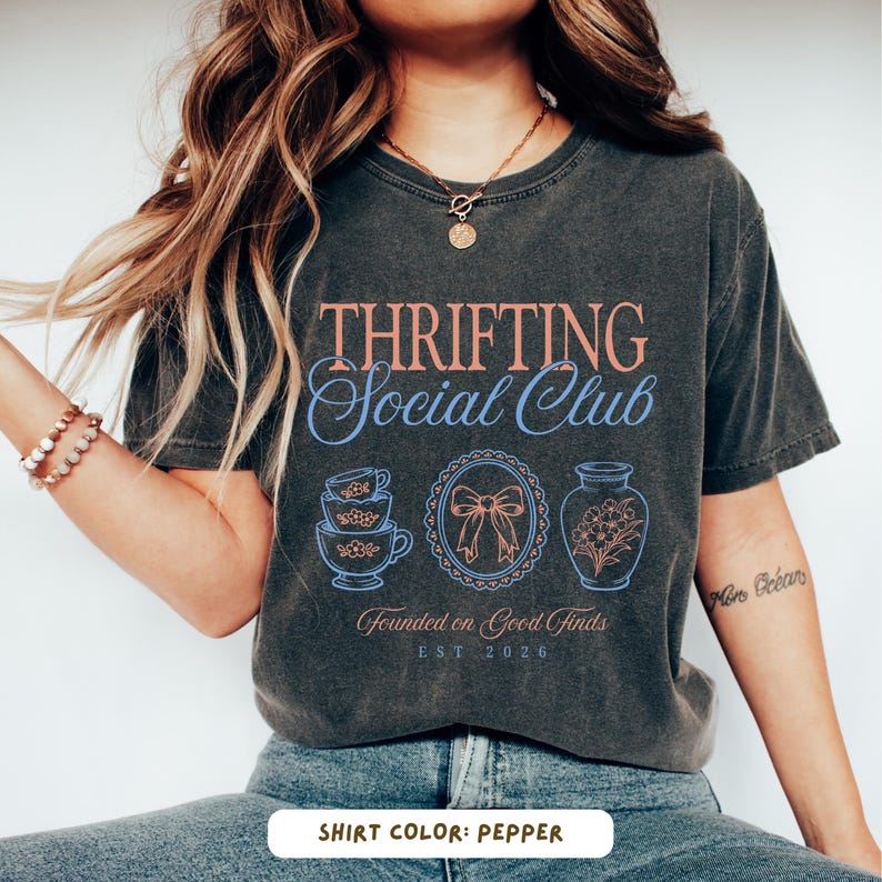 May include: Pepper-colored t-shirt with the text "THRIFTING Social Club" in a vintage style. The design includes illustrations of teacups, a bow, and a vase with flowers. The text "Founded on Good Finds EST 2026" is also included.