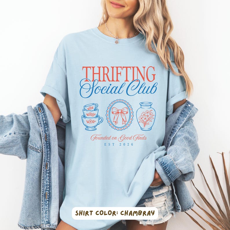 May include: Light blue t-shirt with the text "Thrifting Social Club" in red and blue. The shirt features illustrations of teacups, a bow, and a vase. The text "Founded on Good Finds" and "EST 2026" are also printed on the shirt.