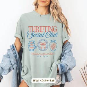 May include: A light green t-shirt with the text "Thrifting Social Club" in red and blue. Below the text are illustrations of teacups, a bow, and a vase. The shirt also has the text "Founded on Good Finds EST 2026".