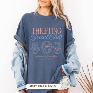 May include: Denim blue t-shirt with the text "Thrifting Social Club" in coral and light blue. The shirt features illustrations of teacups, a bow, and a vase. The text "Founded on Good Finds" and "EST 2026" are also visible.