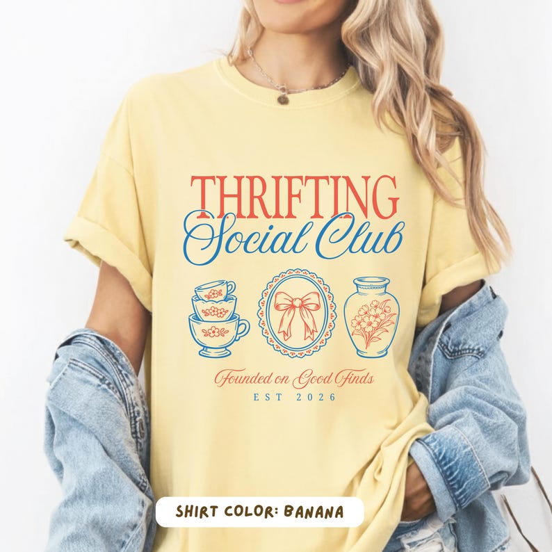 May include: Banana-colored t-shirt with the text "Thrifting Social Club" in red and blue. The design includes illustrations of teacups, a bow, and a vase. The phrase "Founded on Good Finds" and the year "EST 2026" are also present.