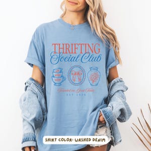 May include: Washed denim blue t-shirt with the text "Thrifting Social Club" in red and blue. The shirt features illustrations of teacups, a bow, and a vase. The text "Founded on Good Finds" and "EST 2026" are also printed on the shirt.