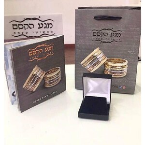 May include: A black jewelry box with a white interior and a black velvet insert. The box is sitting on a white surface. The box is next to a gray paper bag with the text "גני קדם" in Hebrew. The bag is decorated with two gold and silver rings.