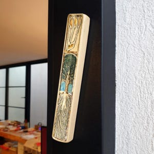 Op de afbeelding: Een keramische mezuzah met een levensboomontwerp. De mezuzah is beige met blauwe, groene en gouden accenten. De mezuzah is gemonteerd op een zwart oppervlak.