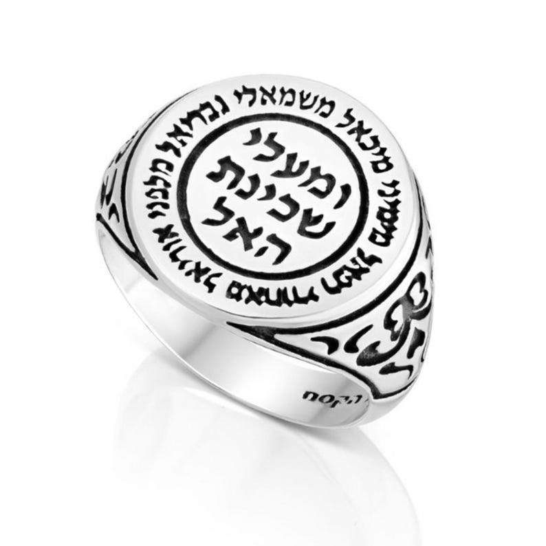 May include: A silver ring with a round, black enamel top featuring Hebrew text. The ring has a decorative, ornate design around the band.