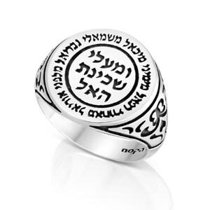 May include: A silver ring with a black inscription in Hebrew. The inscription is in a circle with a smaller circle inside. The ring is engraved with a decorative pattern.