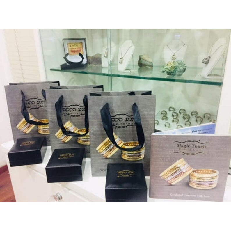 May include: Four gray paper bags with black handles and gold lettering that reads "Magic Touch Jewelry" in Hebrew. The bags are filled with stacks of gold and silver bangles. There are also three black boxes with gold lettering that reads "Magic Touch Jewelry" in Hebrew. The boxes are on a white surface. A brochure with the text "Magic Touch Jewelry" is on the right side of the image.