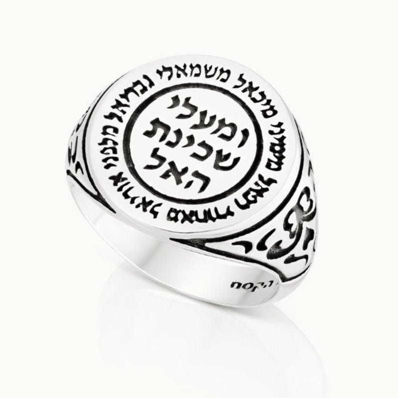 May include: A silver ring with a black inscription in Hebrew. The inscription is surrounded by a circle and the ring is decorated with a floral design.