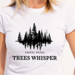 Puede incluir: Camiseta blanca con un gráfico negro de una silueta de bosque y su reflejo. El texto "TRAVEL WHERE TREES WHISPER" está impreso debajo en una fuente negrita sin serifa.