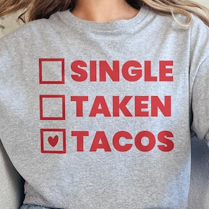 May include: A grey t-shirt with the words "SINGLE", "TAKEN", and "TACOS" printed in red. The word "TACOS" has a heart symbol. The shirt is worn with blue jeans.