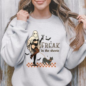 May include: A light gray crewneck sweatshirt featuring a graphic of a ghost wearing lingerie and holding a martini glass, walking a black cat on a leash. The text reads "FREAK in the sheets" above a checkered orange and white pattern with bats.