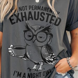 Puede incluir: Camiseta gris con un gráfico de un búho con gafas y una taza de café. El texto de la camiseta dice: "No estoy permanentemente agotado, soy un búho nocturno."