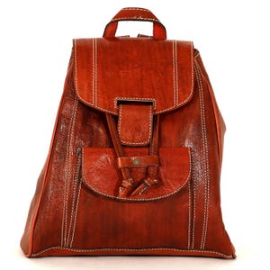 May include: A rust-coloured leather backpack featuring a top flap with a buckle closure, a front pocket with a flap, and a top handle. The bag is accented with white contrast stitching along the edges and seams, providing a structured appearance.