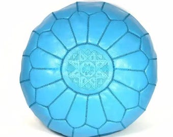 May include: A bright turquoise round leather pouf featuring a central circular panel with intricate geometric embroidery. The surface is divided into petal-shaped segments by visible stitching, creating a structured, symmetrical design.