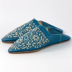 May include: A pair of teal leather slippers featuring a pointed toe and a flat sole. The upper surface of each slipper is decorated with a light beige floral pattern, and the edges are finished with visible light-colored stitching along the seams.