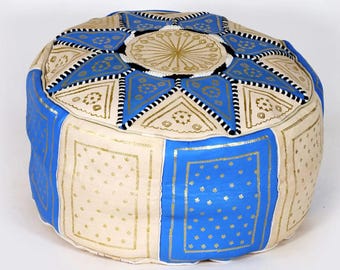 May include: A round floor pouf featuring alternating panels of cream and blue leather. The top surface displays a star-shaped pattern with gold-colored accents and black stitching. The sides are decorated with gold-colored floral motifs.