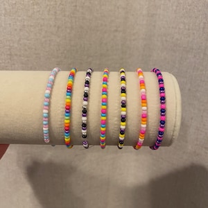 Puede incluir: Siete pulseras de cuentas de colores, cada una con un patrón de color diferente. Las pulseras están dispuestas en una fila sobre una superficie blanca.