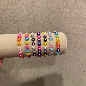Op de afbeelding: Een set van acht kralenarmbanden, elk met een herhalend patroon van kleurrijke bloemvormige kralen. De armbanden zijn in een rij op een wit oppervlak gerangschikt, met kleuren variërend van roze tot oranje, geel, groen, blauw, paars, zwart en wit.