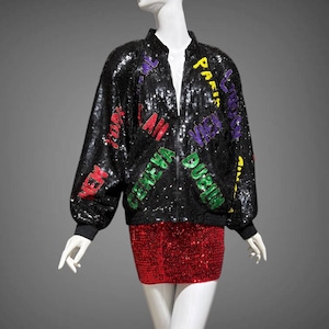 May include: A black sequined bomber jacket with colorful text, paired with a red sequined mini-skirt, displayed on a mannequin. The jacket features text in various colors, including red, green, purple, and yellow. The outfit is a bold fashion statement.