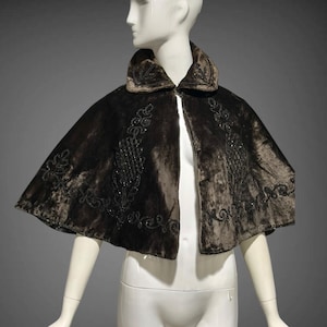 May include: A brown velvet cape with intricate beaded embroidery. The cape features a rounded collar and a flowing, open front design. The embroidery is concentrated around the collar and along the edges of the cape, adding a touch of elegance to the garment.