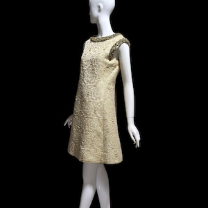 Puede incluir: Vestido vintage de color marfil con un patrón floral texturizado. El vestido sin mangas presenta un escote y detalles en los hombros con cuentas. El vestido se exhibe en un maniquí blanco sobre un fondo negro.