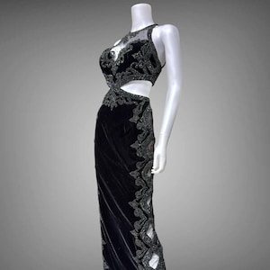 Pode incluir: Vestido de noite preto em um manequim. O vestido apresenta um design recortado na cintura e intrincados enfeites de contas. O vestido tem um decote halter e uma saia longa. O tecido parece ser veludo.