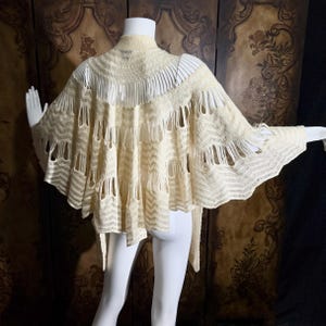 May include: A cream-colored, open-knit cape featuring a high collar and a series of vertical, elongated cutouts arranged in concentric rows. The garment displays a chevron-patterned texture throughout, with a flared, wide silhouette.