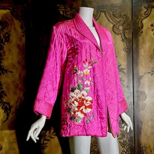 May include: A bright pink quilted jacket featuring a shawl collar and long sleeves. The front is decorated with an intricate embroidered floral design in shades of red, white, and green, showcasing large blooming flowers and delicate stems.