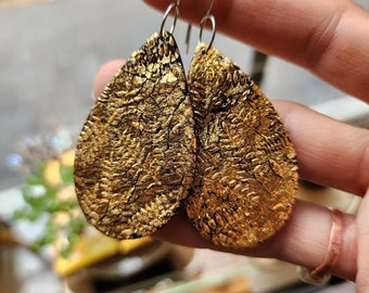 May include: A pair of gold teardrop-shaped earrings with a textured surface. The earrings are made of polymer clay and have silver hooks.