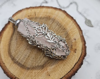 May include: A rose quartz crystal point pendant with a silver chain and floral design. The pendant is resting on a wooden slice.