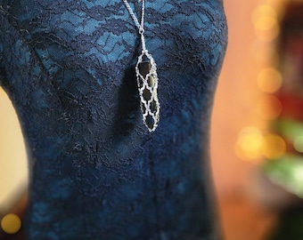 May include: A silver chain necklace with a black pendant featuring a geometric design. The necklace is worn on a mannequin wearing a dark blue lace dress.