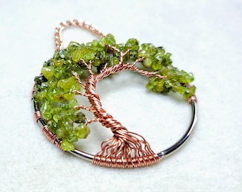 May include: A handcrafted pendant necklace featuring a tree of life design. The tree is made of copper wire with green peridot gemstone leaves. The pendant is set on a silver-colored circular frame and chain.