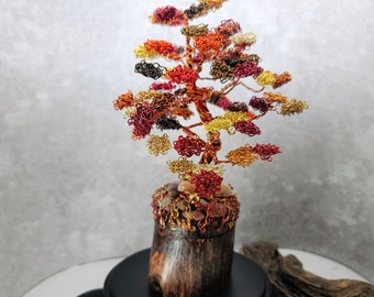 May include: Wire sculpture of a tree with a brown wooden base. The tree is made of copper wire and has a variety of colors, including red, orange, and gold. The tree is on a black base with a wooden texture.
